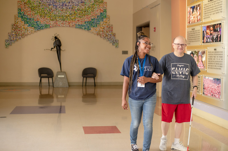 benefits of volunteering Woman walking next to man using a probing cane in a hallway.