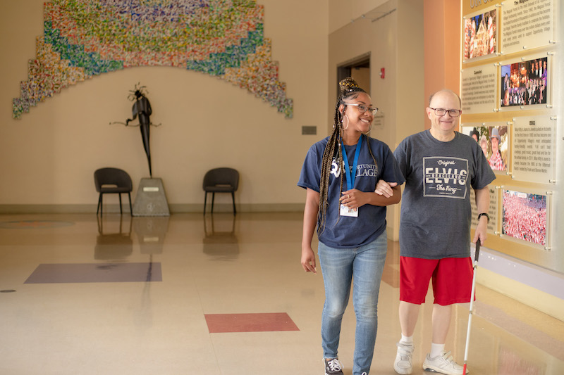 Woman walking next to man using a probing cane in a hallway.