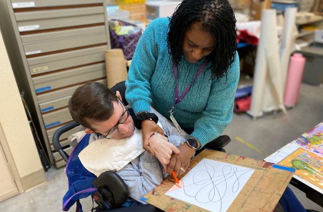 Person helping a person with a disability with drawing on paper.