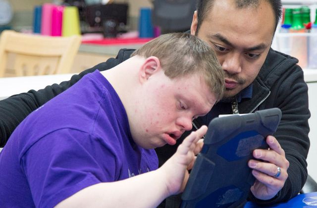 Man helping a young person with a disability use an electronic tablet.