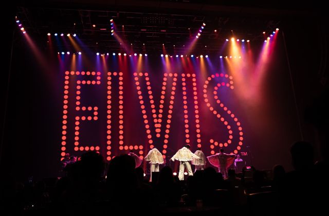 group of people dressed in white suits on stage with their backs towards the audience with a neon sign spelling 'elvis' in the background