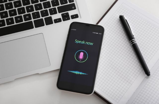 mobile phone displaying the words 'speak now' with a icon of a microphone, setting on a laptop keyboard with a notebook and pen next to it on the table.