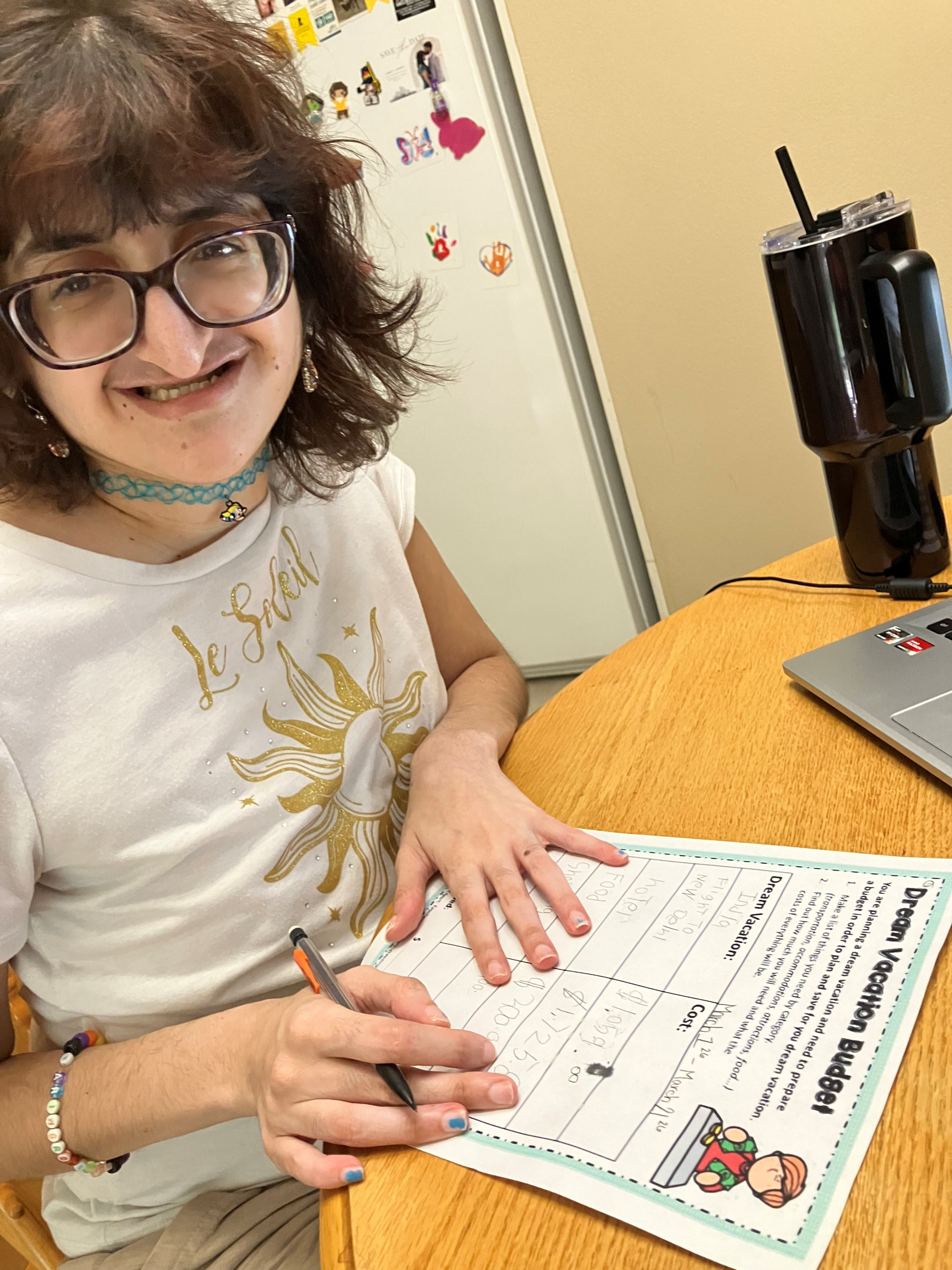 An individual smiles while filling out a vacation budget planning worksheet at a desk during a supported living life skills activity.