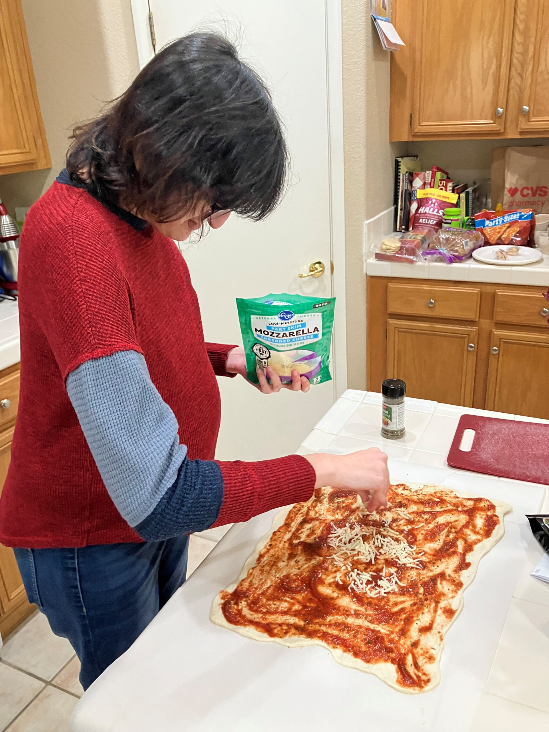 An individual sprinkles shredded mozzarella cheese onto a homemade pizza dough at a kitchen counter during a supported living life skills activity.