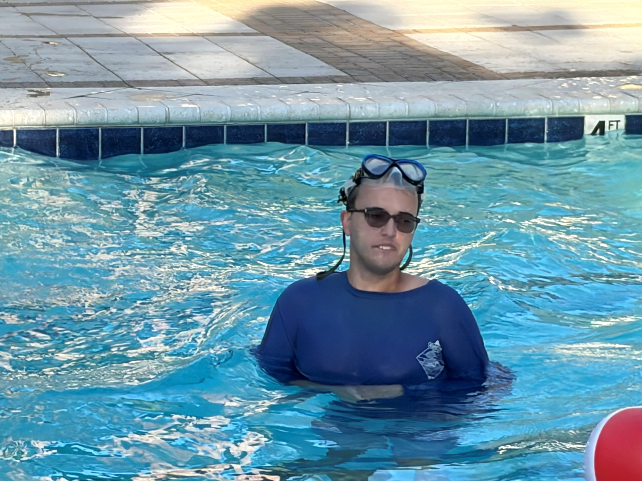 A man wearing a blue swim shirt and a snorkel mask enjoys swimming in an outdoor pool during a supported living activity.