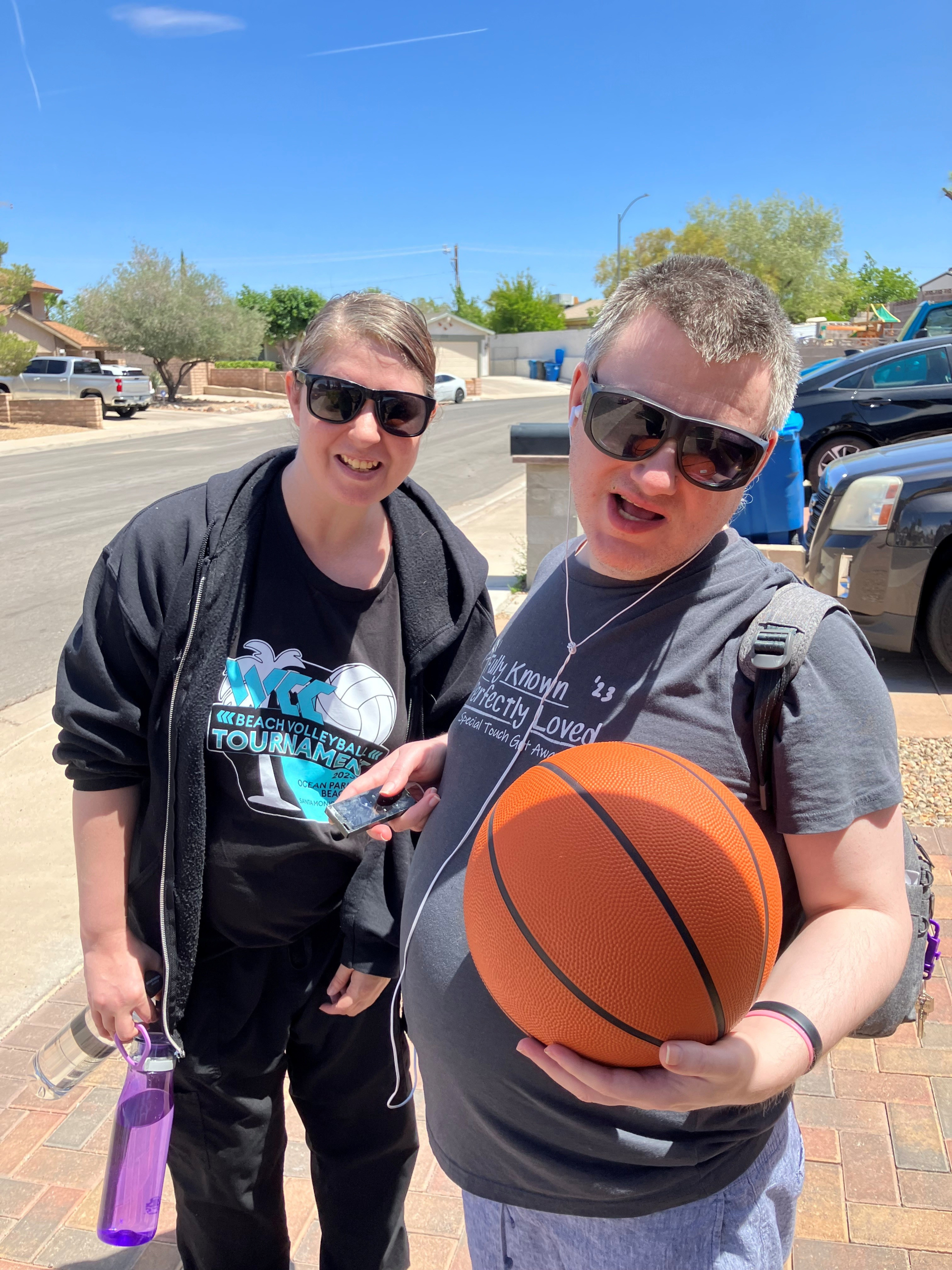Two individuals wearing sunglasses smile together outside on a sunny day while holding a basketball during a supported living activity.