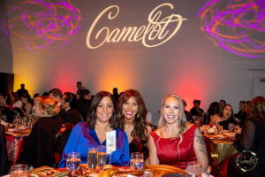 Three women in formal attire smile together at a banquet table during an event with a "Camelot" projection on the illuminated background wall.